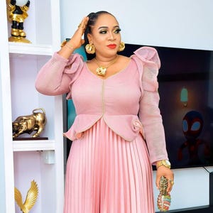 May include: A long, pleated pink dress with a fitted bodice and ruffled sleeves. The dress features a V-neckline and a decorative waist detail. The woman is wearing gold jewelry and holding a decorative clutch.