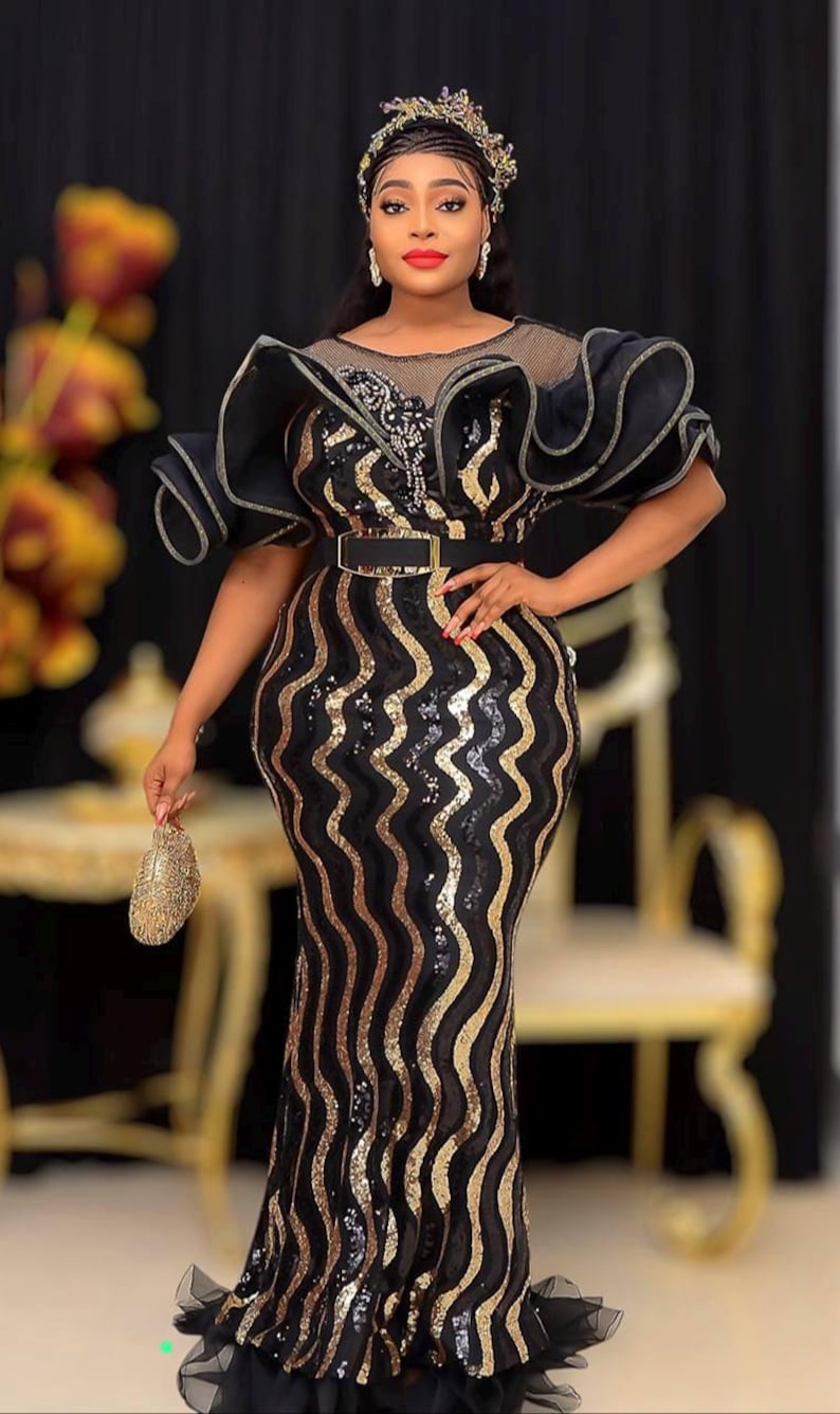 May include: A black and gold sequined evening gown with a wavy stripe pattern. The dress features a black belt, ruffled sleeves, and a small gold clutch. The model is wearing a jeweled headpiece.