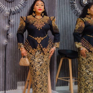 May include: Black and gold formal dress with intricate embroidery. The dress features a fitted bodice with shoulder embellishments and a matching long skirt. A small gold handbag completes the ensemble. The outfit is displayed in a well-lit room with a decorative clock.