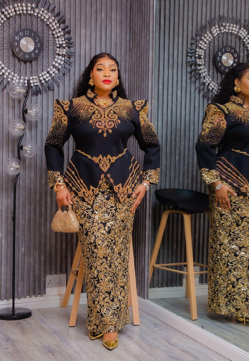 May include: Black and gold formal dress with intricate embroidery. The dress features a fitted bodice with shoulder embellishments and a matching long skirt. A small gold handbag completes the ensemble. The outfit is displayed in a well-lit room with a decorative clock.