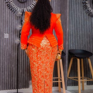 May include: An orange formal dress with gold embroidery. The dress features a fitted bodice with structured shoulders and a peplum waist. The skirt is a long, form-fitting pencil style with a slit in the back. The outfit is accessorized with a small clutch.