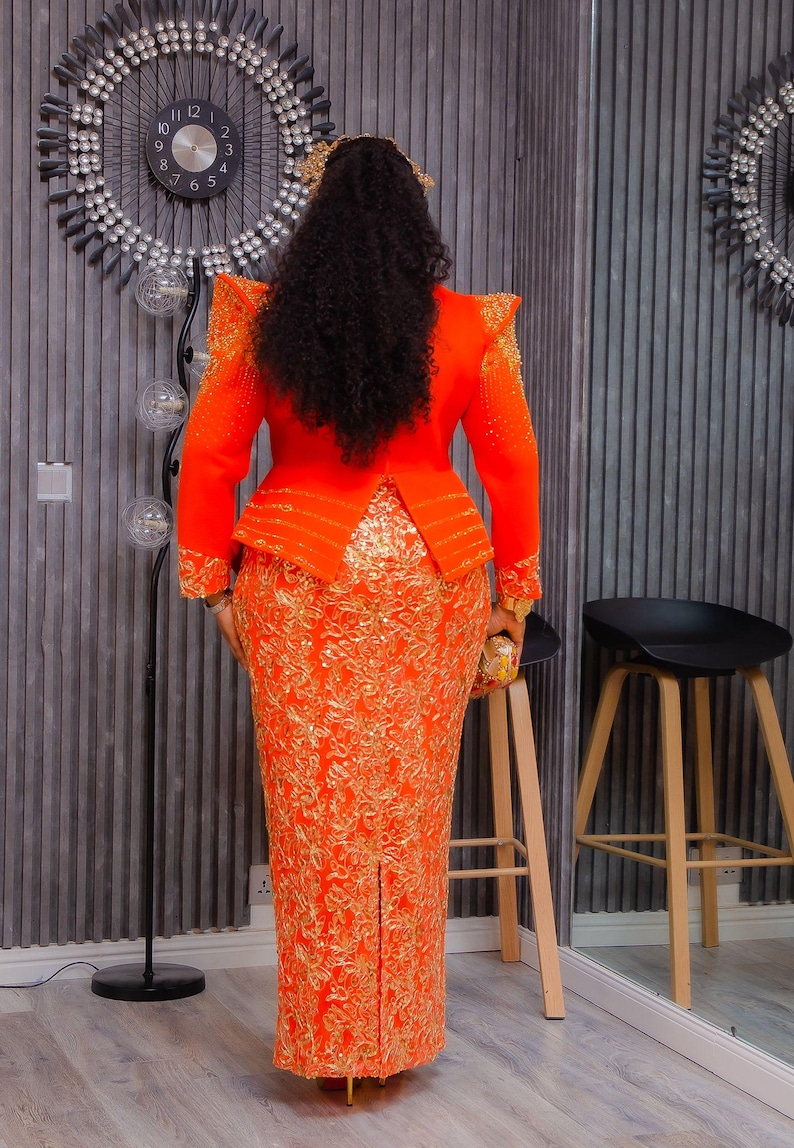 May include: An orange formal dress with gold embroidery. The dress features a fitted bodice with structured shoulders and a peplum waist. The skirt is a long, form-fitting pencil style with a slit in the back. The outfit is accessorized with a small clutch.