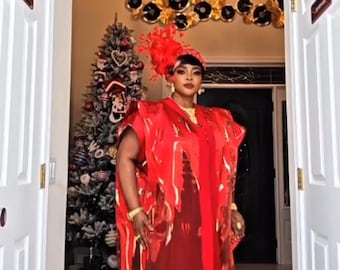 May include: A woman in a red outfit, including a headpiece, cape, and dress, stands between two white doors. A Christmas tree is visible in the background, decorated with ornaments. The outfit has a shiny, transparent overlay.