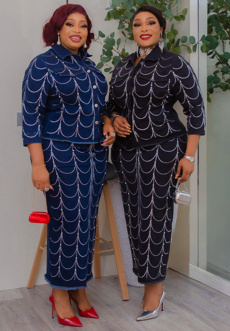 May include: Two women wearing matching denim outfits. One outfit is navy blue, the other is black. Both outfits feature a jacket and skirt or pants, embellished with a silver chain design. The women are wearing heels and carrying small purses.