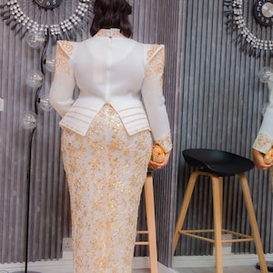 May include: A white and gold formal gown with intricate floral embroidery. The dress features a high neckline, long sleeves, and a peplum waist. Gold embellishments adorn the shoulders and sleeves. The gown is paired with gold heels.