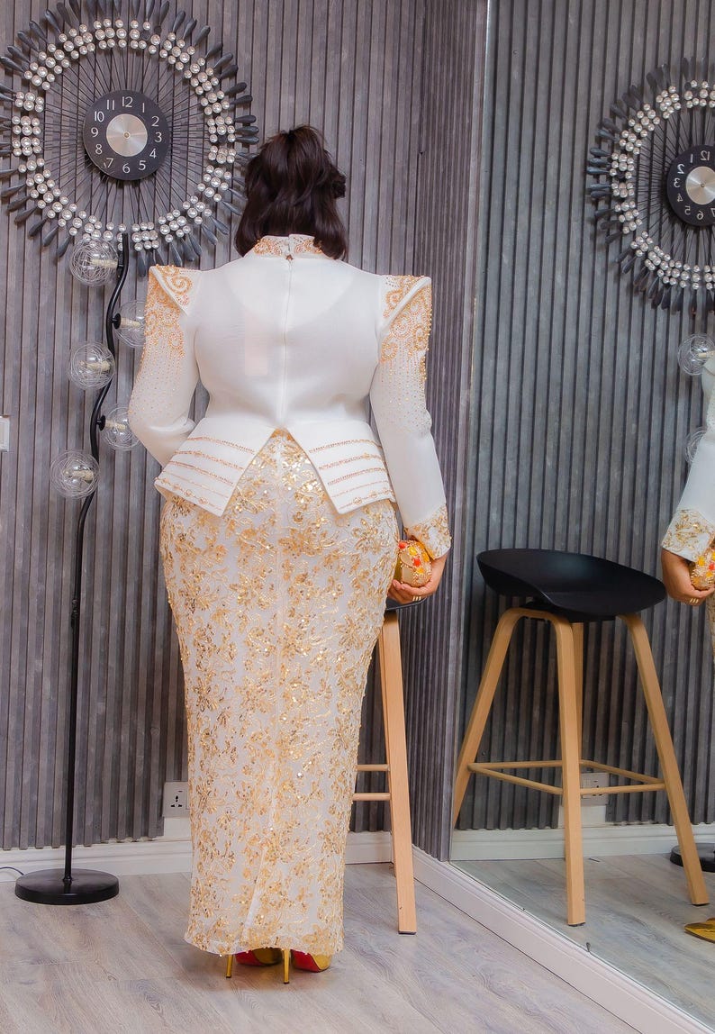 May include: A white and gold formal gown with intricate floral embroidery. The dress features a high neckline, long sleeves, and a peplum waist. Gold embellishments adorn the shoulders and sleeves. The gown is paired with gold heels.