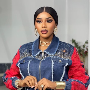 May include: A denim jacket with red lace sleeves and embellishments. The jacket features a zip-up front and is adorned with rhinestones. Gold jewelry, including a necklace, earrings, and bracelets, complements the outfit.