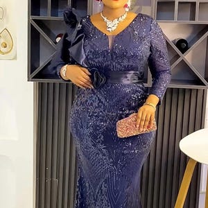 May include: A woman wearing a dark blue sequined gown with a long sleeve and a black belt. She is wearing a pink head wrap with a flower design. She is holding a small clutch purse.
