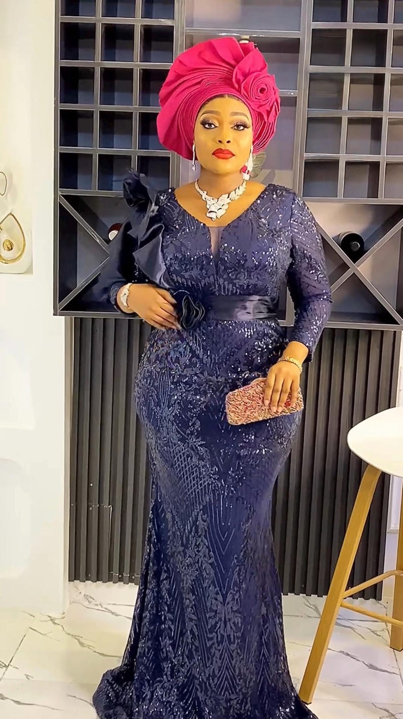 May include: A woman wearing a dark blue sequined gown with a long sleeve and a black belt. She is wearing a pink head wrap with a flower design. She is holding a small clutch purse.