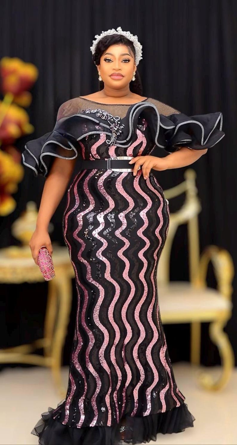 May include: A black and pink sequined formal gown with a wavy pattern. The dress features a sheer neckline, ruffled sleeves, and a silver belt. A matching pink clutch completes the ensemble. The gown is suitable for special occasions.