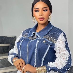 May include: A denim jacket with white lace sleeves and accents, embellished with rhinestones. The jacket has a zipper closure and a frayed collar. The model is wearing gold jewelry, including earrings, bracelets, and rings. The overall style is fashionable and elegant.