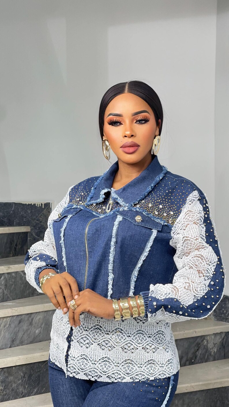 May include: A denim jacket with white lace sleeves and accents, embellished with rhinestones. The jacket has a zipper closure and a frayed collar. The model is wearing gold jewelry, including earrings, bracelets, and rings. The overall style is fashionable and elegant.