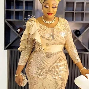 May include: A woman wearing a gold sequined dress with a ruffled shoulder detail and a gold head wrap. She is standing in front of a wine rack.
