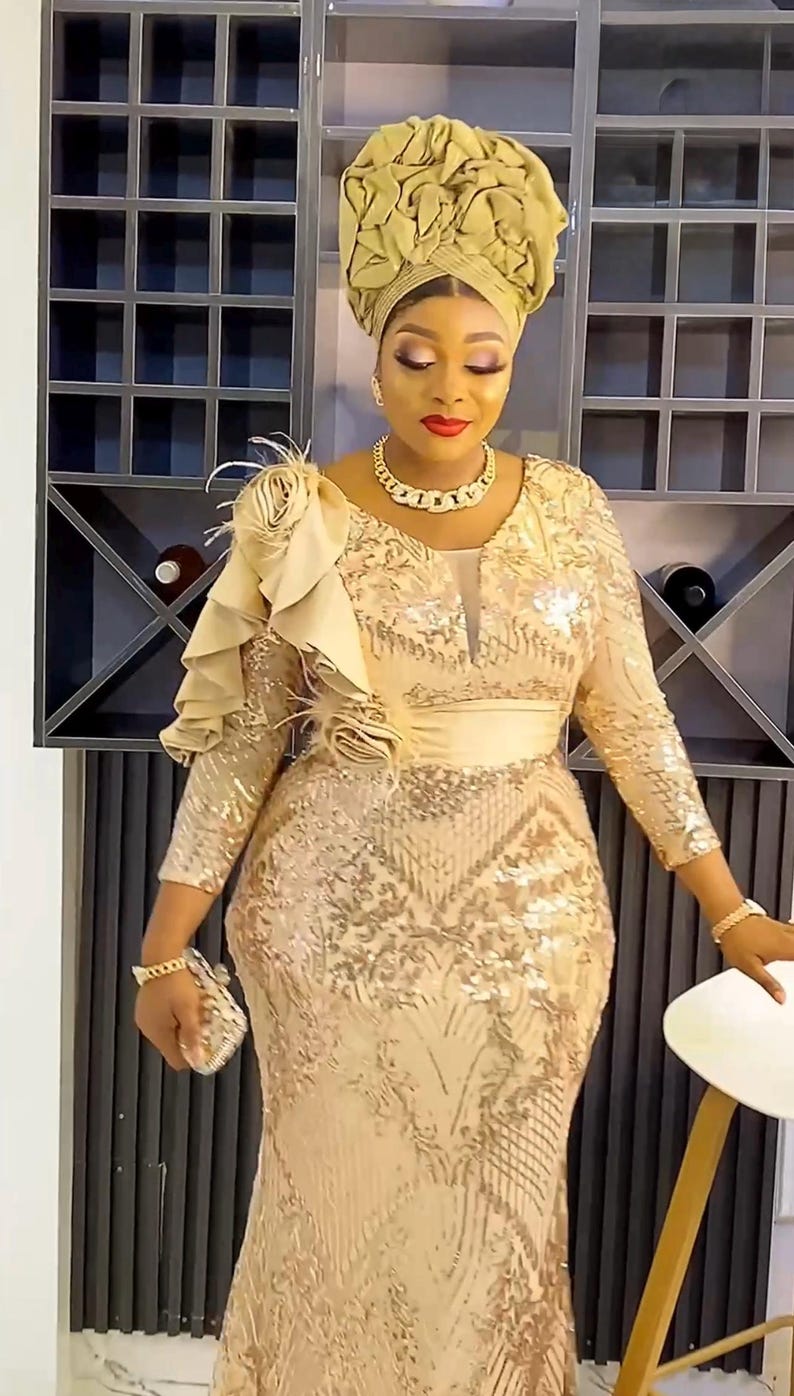 May include: A woman wearing a gold sequined dress with a ruffled shoulder detail and a gold head wrap. She is standing in front of a wine rack.