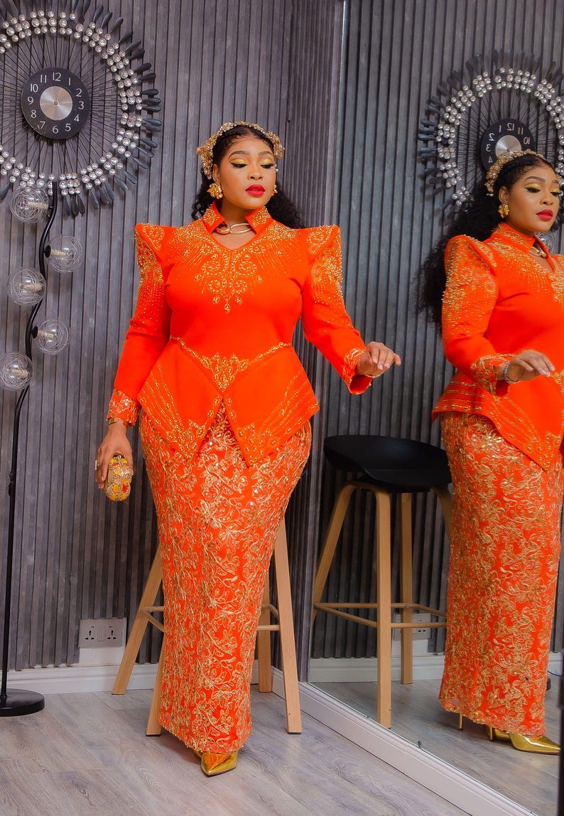 May include: An orange, long-sleeved dress with gold embroidery. The dress features a high collar, structured shoulders, and a fitted waist. The skirt is a matching orange color with gold floral embroidery. A gold clutch and gold heels complete the look.
