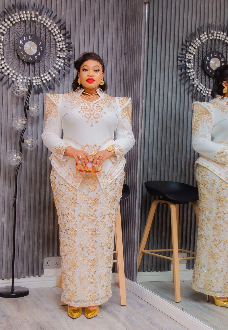 May include: A white formal dress with gold embroidery. The long-sleeved top features shoulder pads and a high neckline. The skirt is a fitted, floor-length design. The outfit is accessorized with gold jewelry and a small clutch.