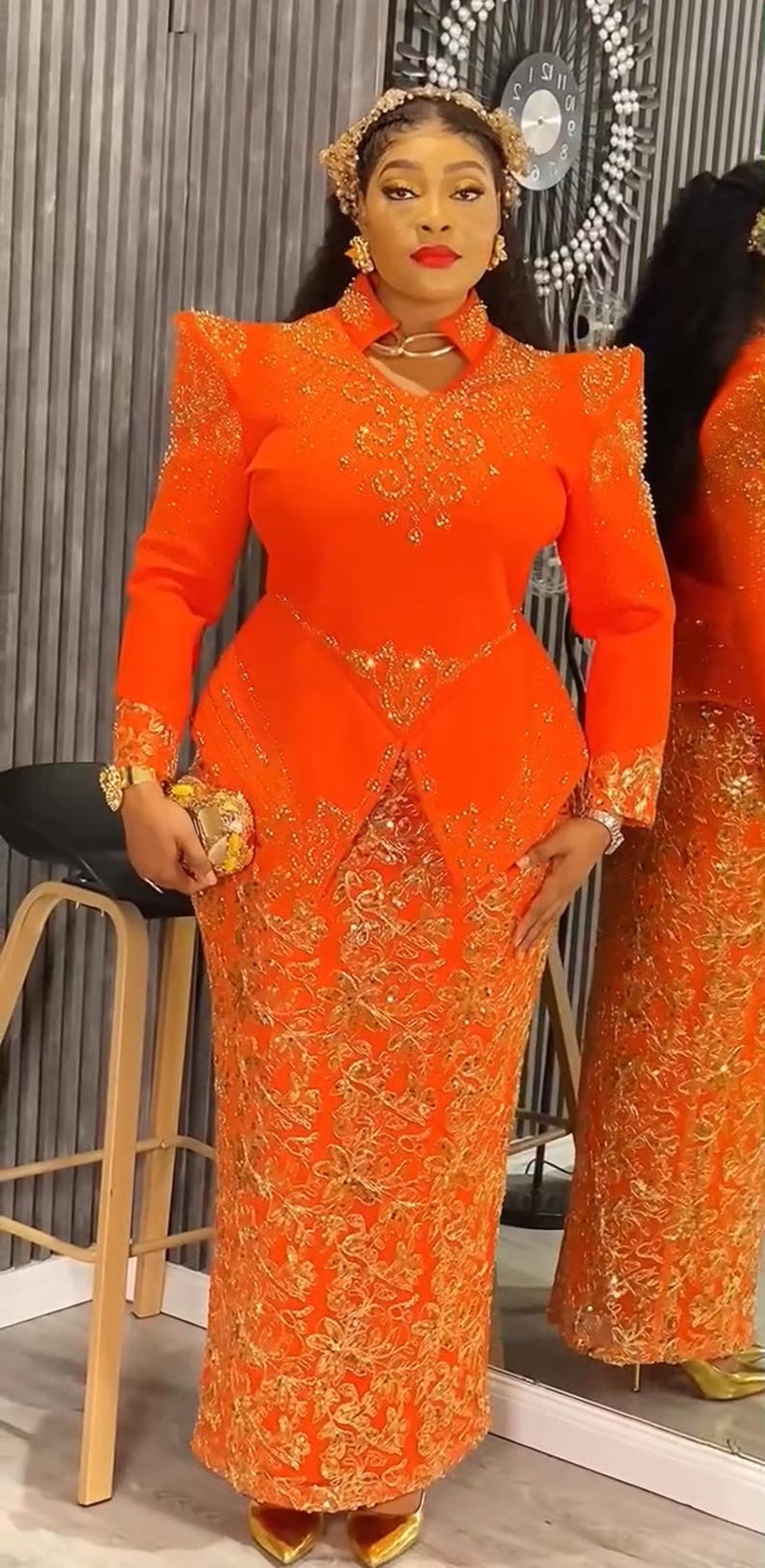 May include: An orange formal gown with gold embroidery. The long-sleeved top has shoulder pads and a high collar. A matching clutch and gold accessories complete the look. The skirt is floor-length.