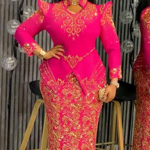 May include: A vibrant fuchsia dress with intricate gold embroidery. The long-sleeved dress features a fitted bodice, a flared skirt, and shoulder embellishments. A gold choker and bracelet complement the outfit. The dress is suitable for formal events.