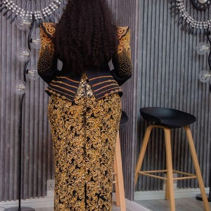 May include: A black and gold formal ensemble. The jacket features gold embellishments on the shoulders and sleeves, with gold accents. The skirt is a matching gold floral pattern. The outfit is worn by a person with long, dark curly hair.