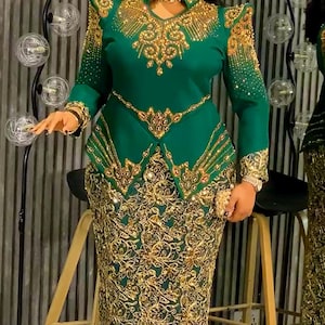 May include: A green formal dress with gold embellishments. The dress features a high neckline, long sleeves, and a fitted bodice with intricate gold detailing. The skirt is a matching green with gold floral patterns.