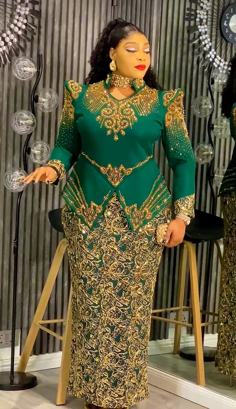 May include: A green formal dress with gold embellishments. The dress features a high neckline, long sleeves, and a fitted bodice with intricate gold detailing. The skirt is a matching green with gold floral patterns.