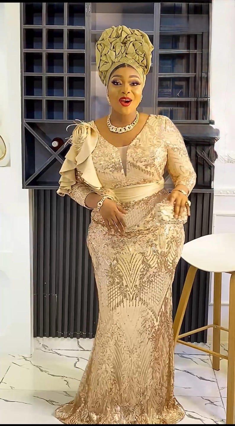 May include: A gold sequined gown with a ruffled shoulder detail and a matching sash. The gown has a fitted bodice and a flowing skirt. The model is wearing a gold head wrap.
