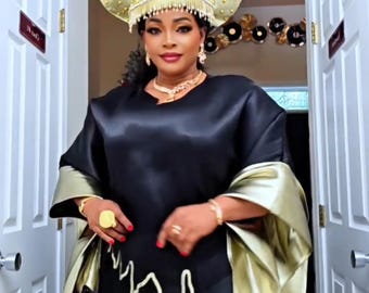May include: A woman wearing a black satin dress with gold accents and a matching headpiece. The dress features gold trim and a flowing design. The headpiece is adorned with gold embellishments and pearls. The outfit is accessorized with gold jewelry.