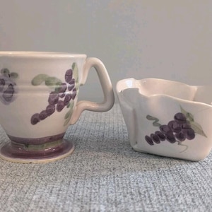 Può includere: Un set di panna e ciotola piccola in ceramica bianca. Entrambi i pezzi presentano un disegno dipinto a mano di uva viola e foglie verdi. La panna ha un manico e un beccuccio. La ciotola ha un bordo ondulato.