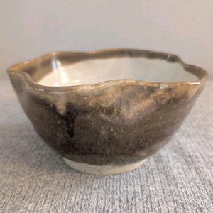 Peut inclure: Un bol en céramique fait à la main, de forme organique unique. L'extérieur présente un glaçage marron moucheté, tandis que l'intérieur est blanc. Le bord est ondulé, ajoutant à son attrait artistique. Parfait pour servir ou comme pièce décorative.