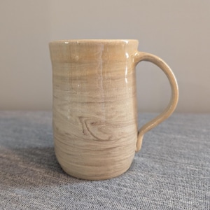 Puede incluir: Una taza de cerámica beige con un patrón de veta de madera. La taza tiene un cuerpo redondeado y un asa.