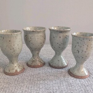 May include: Four handmade ceramic goblets. Each goblet has a speckled, light green glaze with brown spots. The base and rim are a darker brown. The goblets are approximately 10 cm tall.