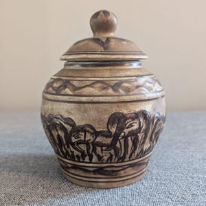 Könnte beinhalten: Eine kleine, keramische Dose mit Deckel, verziert mit einem braunen und weißen Elefantenmuster. Die Dose hat eine braune Glasur und ist handbemalt mit schwarzen Details.