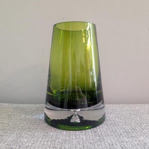 May include: A green glass vase with a flared top and a clear, reflective base. The vase has a smooth, polished surface and a modern, minimalist design. The green colour is vibrant and translucent.