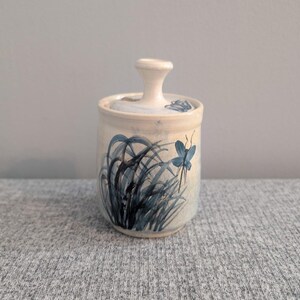 May include: A ceramic jar with a lid, decorated with blue grass and a butterfly design. The jar is cylindrical, with a light beige base and a white lid with a small handle. The design is in shades of blue, suitable for storage.