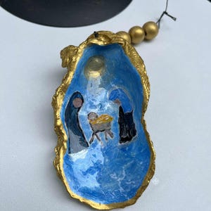 Può includere: Conchiglia di ostrica dipinta a mano con una scena della natività. L'interno è dipinto di blu con un bordo dorato. La scena raffigura Maria, Giuseppe e Gesù bambino in una mangiatoia. Perline dorate sono attaccate per appenderla.