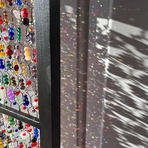 May include: A decorative beaded curtain with a black metal frame. The curtain features a variety of colorful beads in red, blue, yellow, and purple. The sunlight casts a rainbow of colors on the wall.