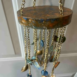 May include: A handcrafted wind chime featuring a circular, aged copper-toned top suspended by gold-colored chains. Decorative elements include small, colorful, shell-like pieces and blue and green beads, creating a unique, artistic design.