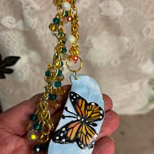 May include: A handcrafted pendant with a painted monarch butterfly design on a light blue, teardrop-shaped stone. It hangs from a gold-coloured chain with colourful beads in green, orange, and clear.