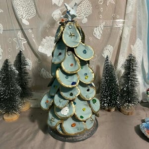 May include: A decorative Christmas tree made of light blue oyster shells with gold trim and colorful gems. The tree is topped with a starfish and sits on a gray base. Small, green bottle brush trees are in the background.
