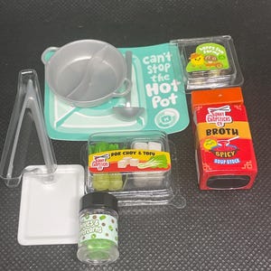 Könnte beinhalten: Ein Miniatur-Hot-Pot-Spielset mit einem silbernen, unterteilten Topf, einer Schöpfkelle, einer transparenten Zange und einem weißen Tablett. Enthält Packungen mit Bok Choy & Tofu, Spicy Soup Stock, Chives & Peppercorns und eine Karte can't stop the Hot Pot.