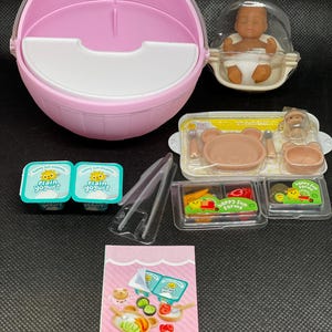 Pode incluir: Uma coleção de comida e acessórios para bebés em miniatura, incluindo um porta-bebés rosa, uma boneca num casulo transparente, iogurtes e um conjunto de pratos em forma de urso. A embalagem apresenta o texto "Happy Sun Farms" e "Yogurt & Fruit".