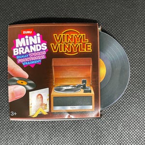 以下が含まれることがあります： Zuru Mini Brands Vinylのおもちゃセット。茶色のミニチュアレコードプレーヤー、黒いターンテーブル、小さな黒いレコードが含まれています。パッケージにはMini Brands Vinylの文字とレコードプレーヤーのグラフィックが印刷されています。