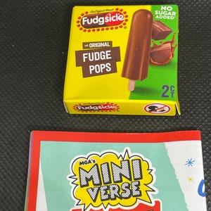 Pode incluir: Uma caixa em miniatura de Fudgsicle fudge pops com um picolé marrom em um palito. A caixa é amarela com as palavras "Fudge Pops" e "No Sugar Added". Abaixo, um cartão com as palavras "MGA's Mini Verse Real Brands".