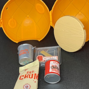 Pode incluir: Um recipiente de plástico amarelo aberto com uma tampa redonda de madeira e vários alimentos em miniatura. Inclui uma lata de "RAW CHUM", um saco de "DRU CHUM" e um pequeno balde de metal.