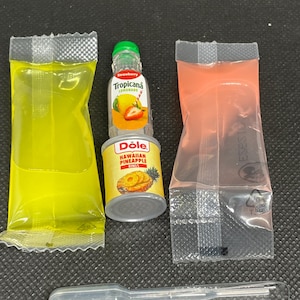 May include: A miniature Tropicana Strawberry Lemonade bottle stacked on a Dole Hawaiian Pineapple Rings can, flanked by two sealed plastic packets containing yellow and pink liquid, and a clear plastic dropper tool positioned below.