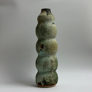 May include: A tall, textured ceramic vase with a unique, stacked design. The vase has a speckled, mottled finish in shades of green, brown, and tan. The opening is dark brown, and the base is a lighter brown. A decorative piece for the home.