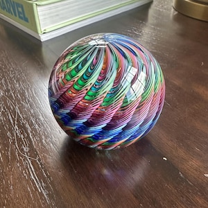 May include: A colorful glass paperweight with a swirling pattern of blue, green, pink, and red. The spherical object reflects light, creating a vibrant display of colors. The decorative item sits on a wooden surface.
