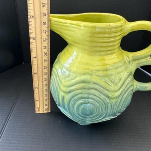 May include: A ceramic pitcher with a yellow-green glaze, transitioning to a teal-green at the base. The pitcher has a spout, two handles, and a textured, concentric circle design. The pitcher is approximately 6 inches tall.