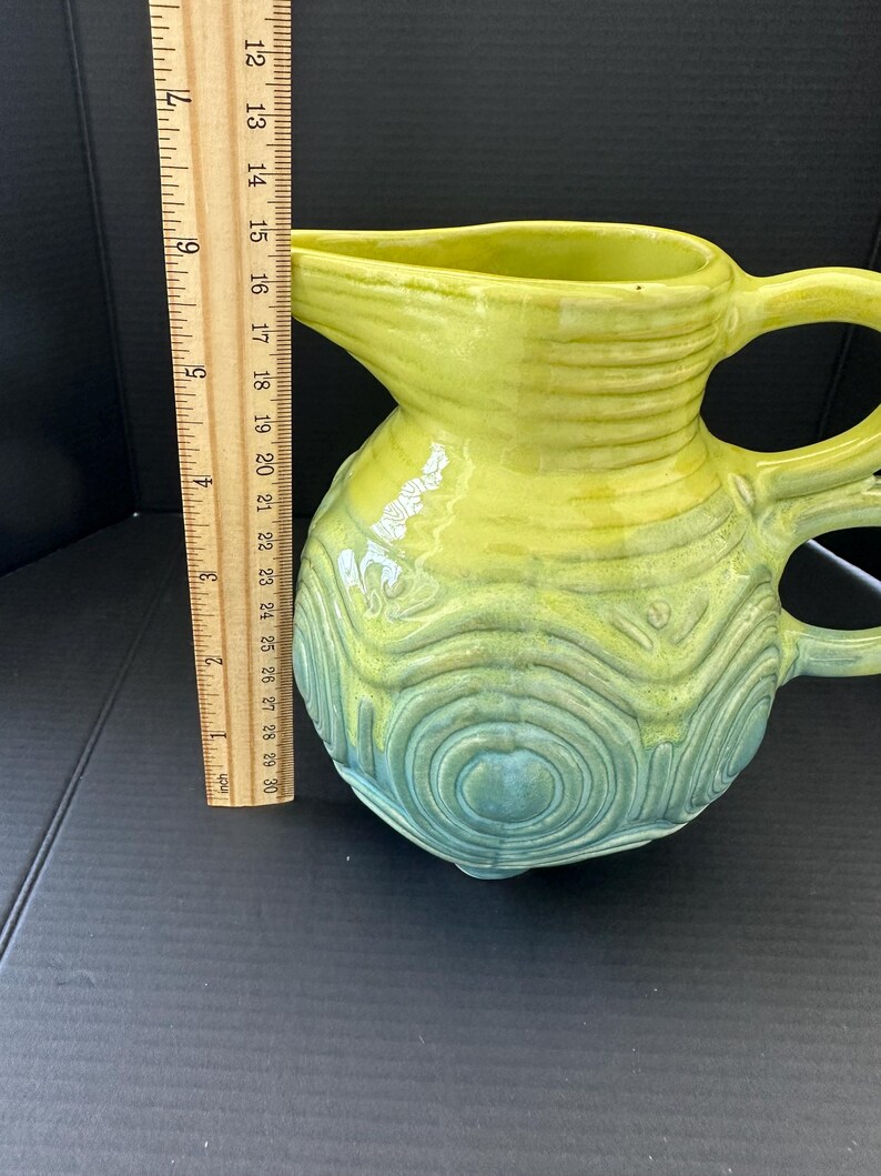May include: A ceramic pitcher with a yellow-green glaze, transitioning to a teal-green at the base. The pitcher has a spout, two handles, and a textured, concentric circle design. The pitcher is approximately 6 inches tall.