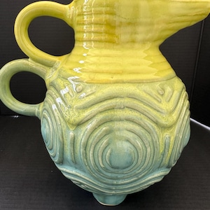 May include: A handcrafted ceramic pitcher with a yellow top and a blue-green base. The pitcher features a spout, two handles, and a textured, concentric circle design. The pitcher is approximately 9 inches tall.